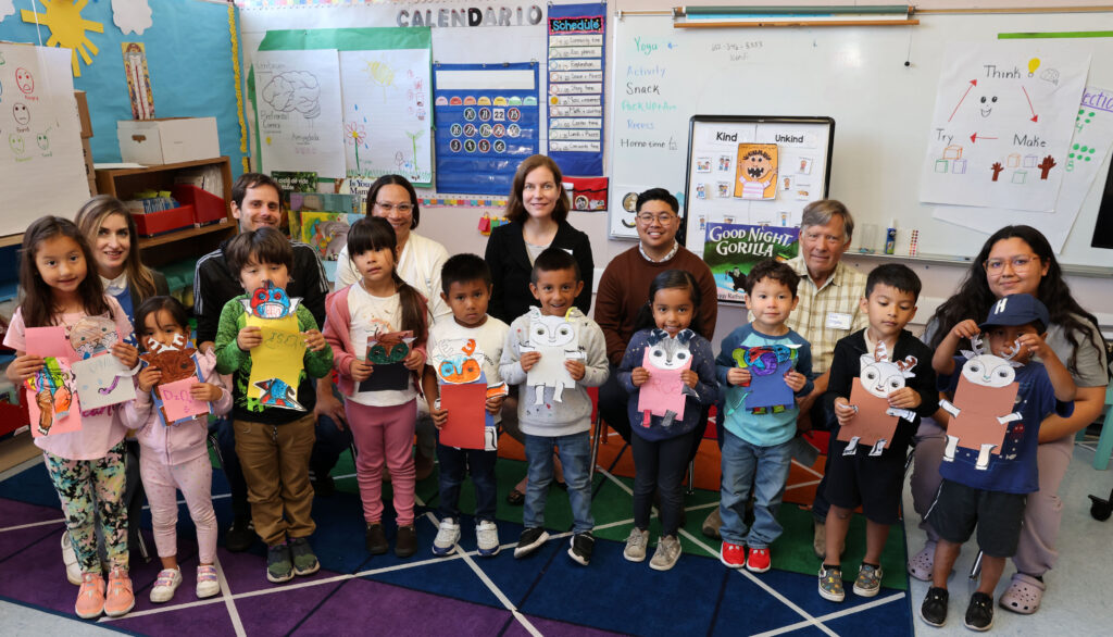 A group of young children holding artwork stands in a row in a classroom with adults kneeling behind them