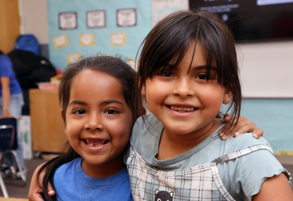 Two elementary school-aged girls smile at the camera with their arms around each other